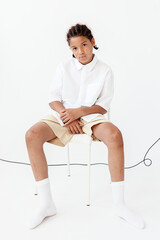 Stylish young boy sitting crosslegged on chair in white shirt and shorts, exuding trendy vibes