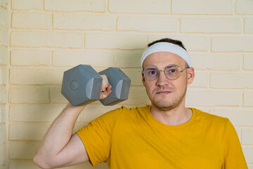 A man wearing a yellow t-shirt, white headband, and glasses lifts a grey dumbbell with his right arm