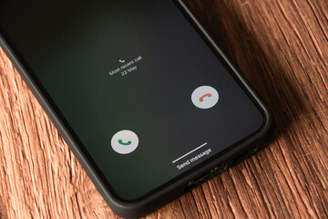 A black phone with incoming call screen with green accept and red deny button on wooden background. Including Send message and Most recent call text. © Laura