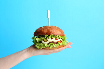 Woman holding delicious vegetarian burger on light blue background, closeup