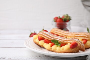 Delicious eclairs filled with cream, strawberries and mint on white wooden table, closeup