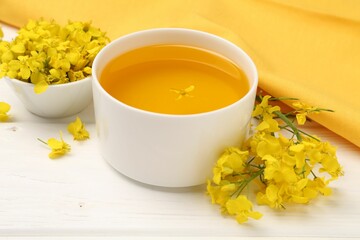 Rapeseed oil in bowl and beautiful yellow flowers on white wooden table