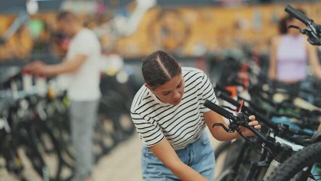 Young female shopper carefully inspecting mountain bicycle handlebars and controls in store, showing curiosity and determination in search for right bike for adventures