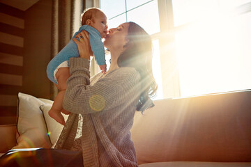 Mother, baby and kiss on cheek in living room on sofa with love for relationship with new born, care. Embrace, light and sunshine for relax on couch with playing, joy and happiness for wellness.