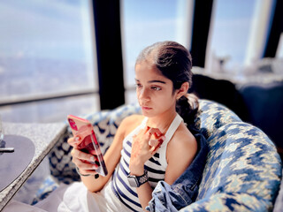 A thoughtful young girl pausing as she uses her smartphone in a sunlit, high-rise lounge with a cityscape visible through the window.