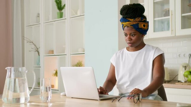 African American girl sitting at kitchen table with laptop working remote as freelancer or student. Black skinned woman gets bad news or setback. She looks upset and angry, shouting and gesturing
