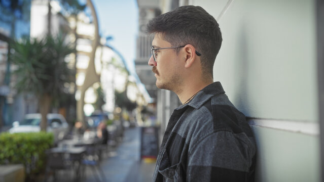 Profile view of a young hispanic man with glasses and a moustache, standing on a city street with urban background.