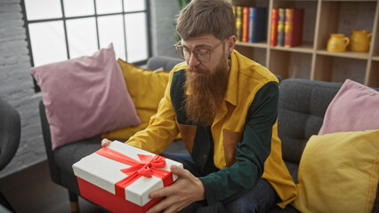 Bearded man with glasses contemplating a gift box indoors on a cozy sofa surrounded by colorful...