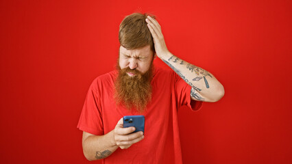 Stressed out young redhead guy, smartphone in hand, looking upset over disturbing message, standing...