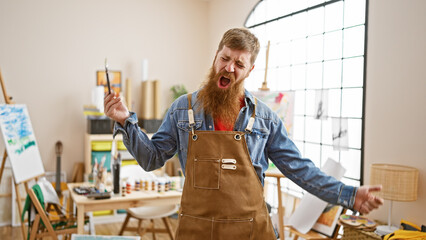 Upbeat, young redhead artist guy, joyfully swinging his paintbrush to the music in art studio,...