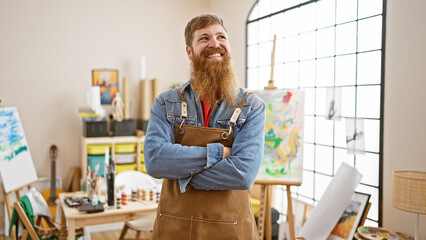 Bearded handsome redhead man, young artist, stands confidently in art studio with arms crossed, smiling brightly while enjoying his painting class