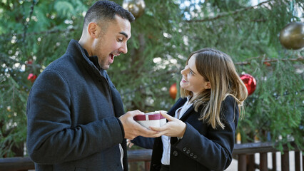 Surprised man receiving gift from woman outdoors by christmas tree