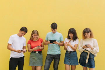 Group of teenagers using their mobile phones and tablet on a yellow background with copy space