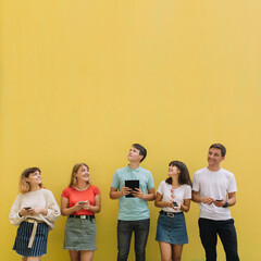 Group of teenagers using their mobile phones and tablet on a yellow background with copy space
