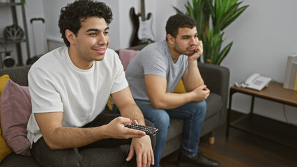 Two hispanic men relaxing indoors, one holding a remote and smiling, the other contemplative and serious
