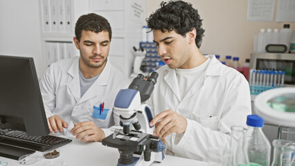 Two male scientists working together in a laboratory, using a microscope and computer for research.