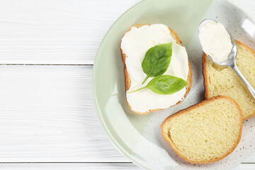 Pieces of bread with cream cheese and basil leaves on white wooden table, top view. Space for text