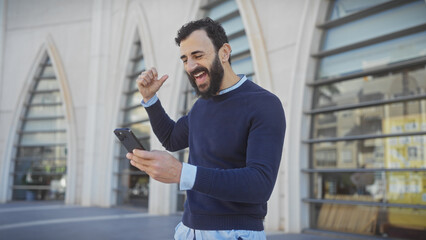 Cheerful bearded man celebrating success while using a smartphone on a city street.