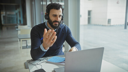 Bearded man in headphones gesturing during a video call in a modern office setting.
