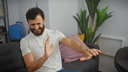 Fototapeta premium A bearded man winces in pain, holding his arm inside a modern living room, conveying injury or discomfort.