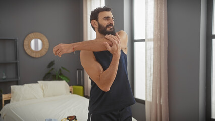 A bearded man stretching his arms in a modern bedroom setting, exhibiting morning fitness routine...