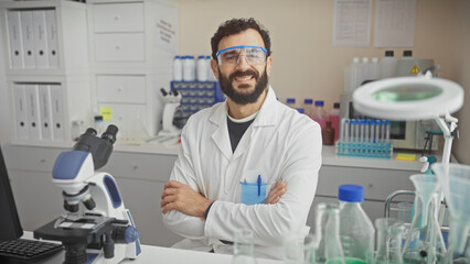 Smiling bearded man in lab coat, safety goggles, standing in lab with microscope and scientific...