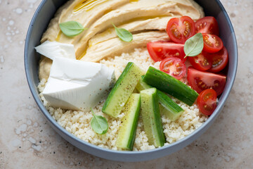 Grey bowl with hummus, couscous, feta cheese, tomatoes and cucumber, horizontal shot, middle close-up