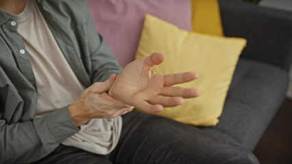 Hispanic man with beard in pain holding wrist in modern living room setting, expressing discomfort and health issues.