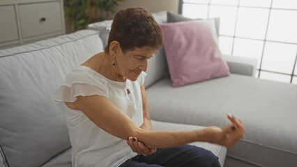 Obraz premium An elderly hispanic woman with short hair massages her elbow while sitting on the couch in a cozy living room, suggesting joint or muscle pain in an indoor home setting.