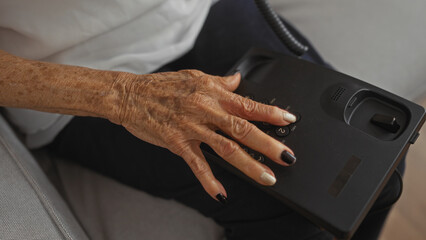 An elderly woman using a black landline phone in her living room at home, with her mature hands showcasing age marks and a neatly polished manicure.