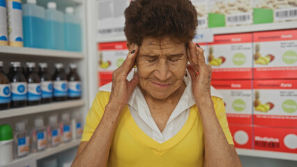 An elderly hispanic woman with short hair wearing a yellow shirt holds her head in pain while standing in a pharmacy surrounded by various medications.