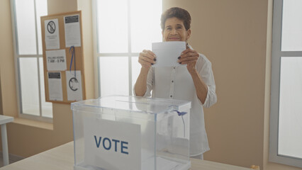 A senior hispanic woman with short hair votes in an indoor electoral room, placing her ballot in a...