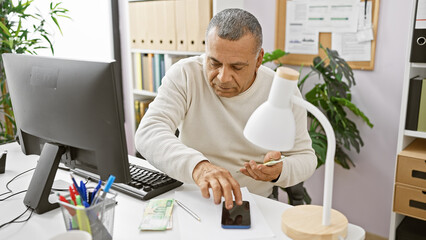 Mature hispanic man working in office using smartphone, money, and computer on desk.