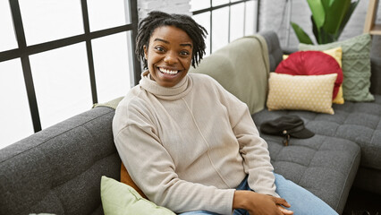 Smiling young black woman with dreadlocks sitting on a sofa in a cozy living room setting, portraying a relaxed and comfortable atmosphere.