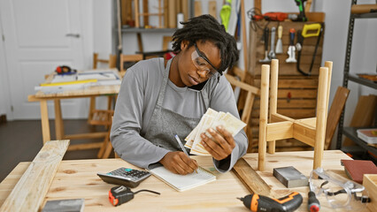 African woman in workshop counting danish currency while talking on phone surrounded by woodworking tools.