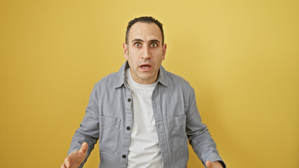 A shocked hispanic man in casual attire standing against a yellow isolated background, expresses surprise with wide eyes and an open mouth.