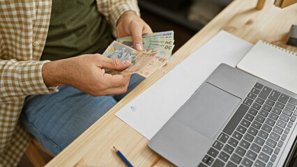 A young man counts romanian currency at a workshop indoors with a laptop and notepad on the desk.