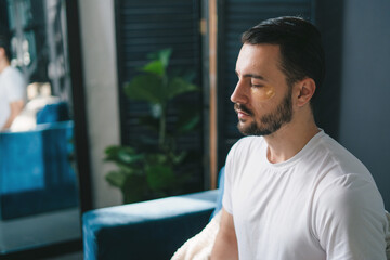 Handsome man with eyes closed and eye-patches sitting on couch in lotus pose and meditating at home.