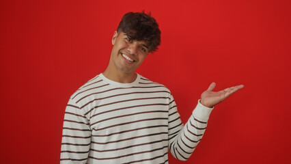 Fototapeta premium Handsome young hispanic man presents with an extended hand against an isolated red background.