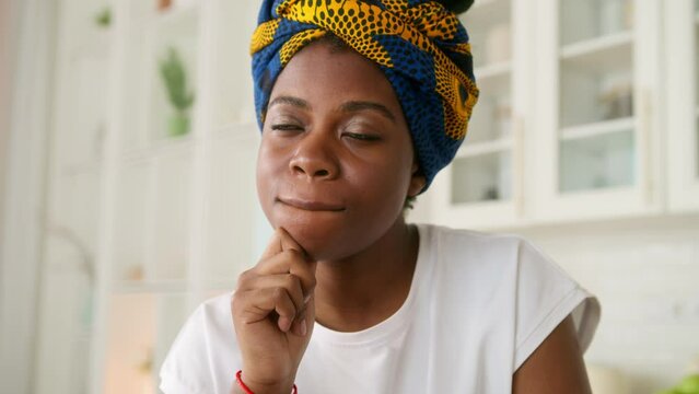 Pensive African American girl sitting at kitchen table with laptop. Young dark skinned woman thinks over work or daily issues, looking through window focused and deep in thoughts