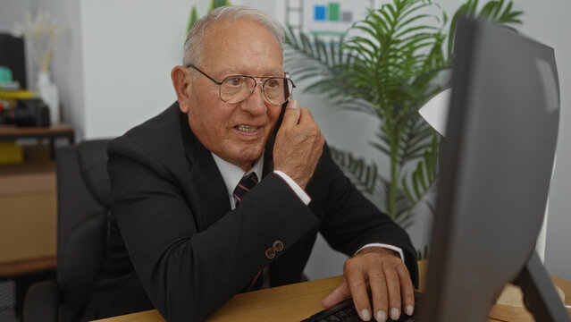 Elderly, grey-haired, caucasian man in a suit, speaking on a mobile phone, sits in a modern office, with indoor plants and office supplies visible in the background.