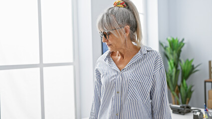 Mature woman with grey hair in a striped shirt stands thoughtfully in a bright office setting, embodying professionalism and experience.