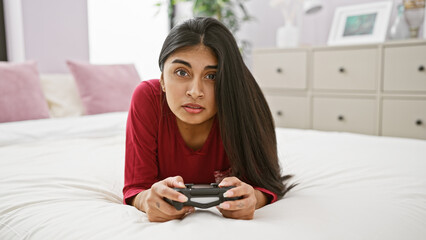 Focused indian woman playing a video game in a well-lit bedroom, lying on a white bed