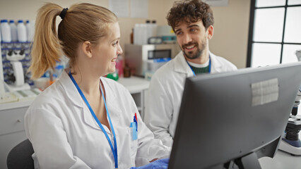 Man and woman in lab coats smiling at each other by a computer in a bright laboratory setting.