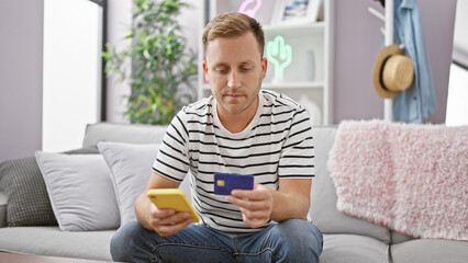 Handsome young caucasian man relaxed, sitting on his living room sofa, engaging in online shopping using his smartphone, texting payment details with a credit card at home.