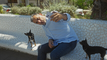 Middle-aged hispanic man takes a selfie with two chihuahua dogs on a white mosaic bench in an urban park.