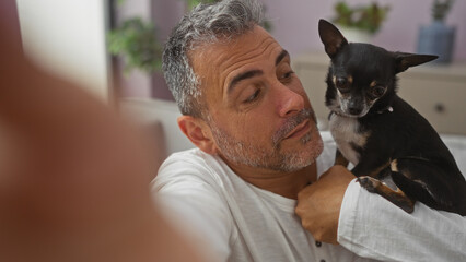 Middle-aged hispanic man holding chihuahua indoors at home, looking at the small dog in a cozy apartment living room.