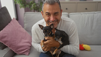 Middle-aged hispanic man holding two chihuahuas while sitting on a couch in a cozy living room.