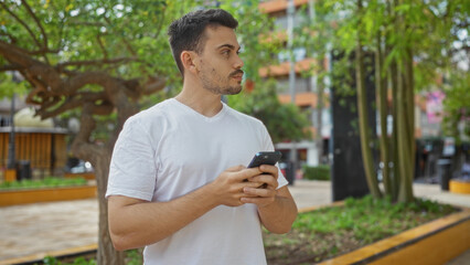 Fototapeta premium A handsome hispanic man with a beard standing in an urban park while holding a smartphone on a bright day outdoors.