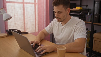 A young, handsome hispanic man working on a laptop in an office setting with a cup of coffee on the...
